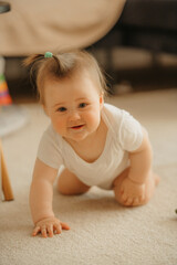 A 7-month girl is kneeling and holding with one hand on the carpet at home. A curious infant in a bodysuit in the sunlight.