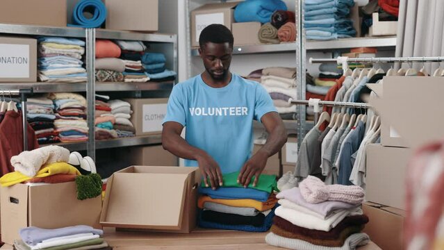 African american man standing at the warehouse while sorting and iterating clothes for donations. Belongings at the shelves at the background. Humanitarian aid concept