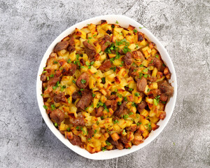 Oven baked meat with potatoes and herbs in a white baking dish on a dark grey background. Top view, flat lay