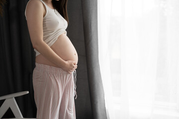 Pregnant woman holding her hands on her belly near the window at home.Maternity, pregnancy, happiness concept