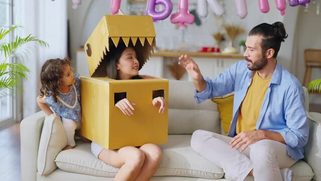 Happy family of parents and daughter child girl playing dinosaurs hunt at home, dad wearing handmade costume monster of cardboard chasing mom and daughter with fun and joyful exited in living room