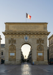 Fototapeta na wymiar Early summer morning view of ancient Arc de Triomphe or triumphal arch historic monument with French flag, famous landmark of Montpellier, France