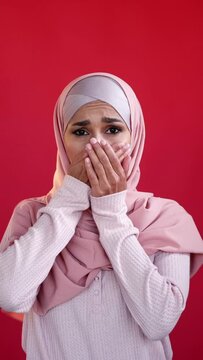 Vertical video. Omg shock. Regret sorrow. Grief compassion. Portrait of overwhelmed sad upset woman in hijab covering mouth with hands on red.