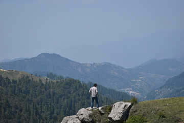 Traveler guy standing on cliff side rock and beautiful cedar tree forest, mountain in background