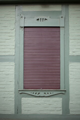 Fenster mit braunen heruntergezogenen Rolläden an einer grünen Hauswand mit Fachwerkbalken,...