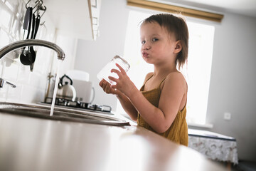 Cute toddler baby washing glass in bright real kitchen