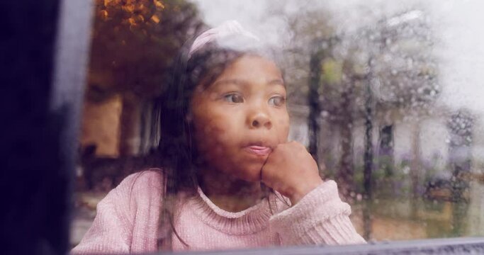 Bored female child looking outside on a wet winter day. Unhappy child feeling lonely and depressed while looking at the bad weather outdoors. Sad little girl standing by the window on a rainy day.
