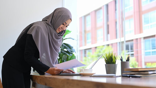 Professional young muslim business woman in hijab standing at office desk and checking financial data on her laptop computer