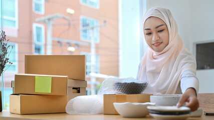 Beautiful muslim woman in hijab preparing product for shipping to customers, working in modern home office
