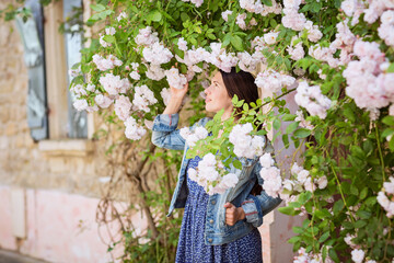 Lifestyle portrait of young stylish woman staying on the street in old town and touching the flowers of white roses, wearing blue dress and denim jacket