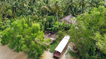 man standing on dock with boat on tropical coastline of Ben Tre along brown Mekong River with coconut tree jungle, aerial