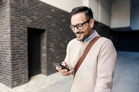 A Happy Smart Casual Man Scrolling On The Phone Outside And Smiling At It.