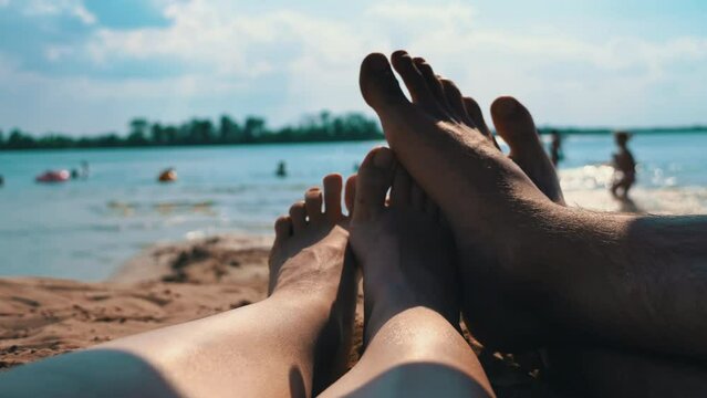 Legs of a couple in love bask on the beach against the background of water. Bare feet of a man and a woman in a romantic sea setting.