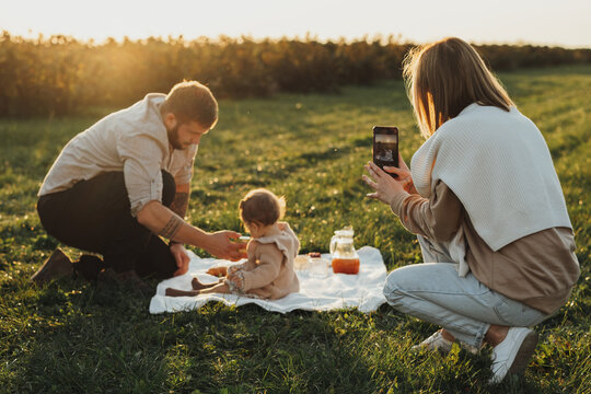 Happy Young Family Having Picnic Outdoors, Woman Taking Pictures of Her Husband and Baby Daughter at Sunset