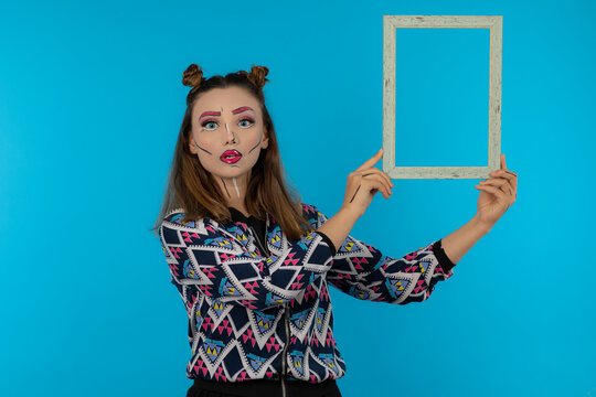 Portrait of attractive girl with creative makeup holding empty frame on blue background