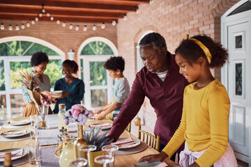 Happy black little girl and her grandfather preparing dining table for family meal on patio.