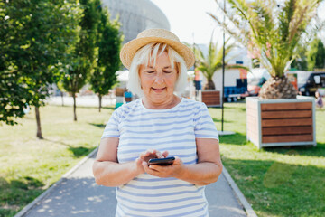 Mature blonde woman in casual summer clothes with straw hat browsing phone while wandering streets of city. Caucasian blogger holding phone pondering thinking on idea for social publication outdoors