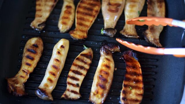 Grilling eringi mushrooms in soy sauce glaze with garlic and herbs on grill skillet in the kitchen. Turning the slices with golden crust. Cooking king oyster mushroom