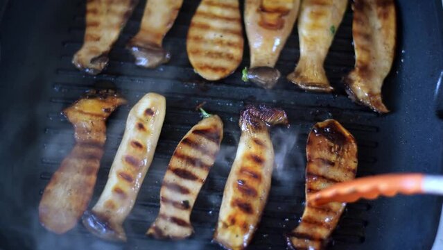 Grilling eringi mushrooms in soy sauce glaze with garlic and herbs on grill skillet in the kitchen. Turning the slices with golden crust. Cooking king oyster mushroom