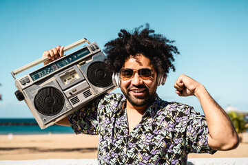 Happy Latin man having fun listening to music with wireless headphones and vintage boombox on the...