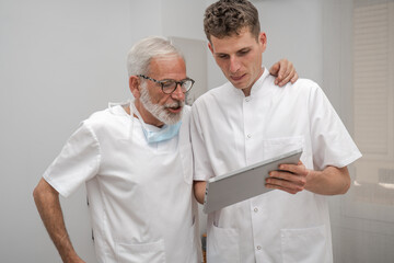 Mature smiling doctor and young intern doctor discussing patient diagnosis, holding digital tablet. Father and son dentist doctors