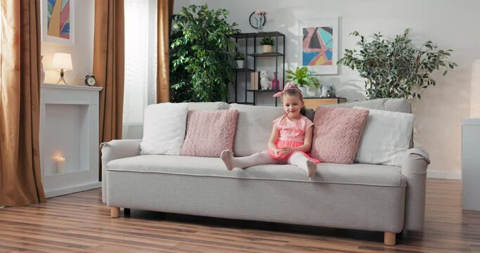 Happy little girl in pink dress with headband is sitting on sofa waving legs smiling happily resting on sofa.