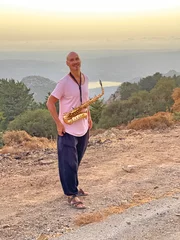Gordijnen Young handsome and bald male saxophonist stands against the sunset with a golden saxophone musical instrument and smiles. In a pink T-shirt and black pants. Musician with a saxophone in nature © Ekaterina