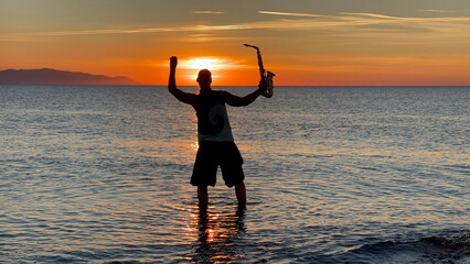 Young male saxophonist stands with his feet in sea water, holds saxophone in his hands, looks at sunset. Beautiful sunset on sea, sky. Musician, playing saxophone, dancing, having fun, Silhouette