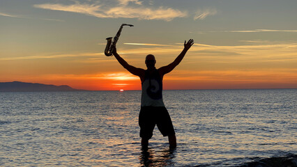 Fototapeta premium Young male saxophonist stands with his feet in sea water, holds saxophone in his hands, looks at sunset. Beautiful sunset on sea, sky. Musician, playing saxophone, dancing, having fun, Silhouette