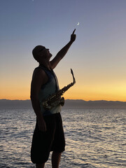 Young male saxophonist stands with his feet in sea water, holds saxophone in his hands. Shows the moon in the sky. Beautiful sunset on sea, sky. Musician, saxophone. Wearing dark black glasses