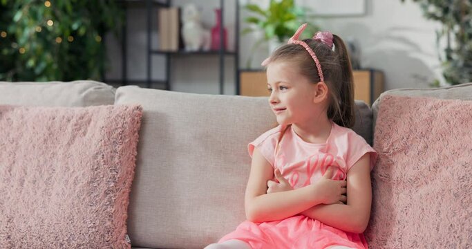 A little girl in a pink dress with a headband is sitting on a sofa, waving her legs, smiling, resting on the sofa looking out the window and then at the camera laughing.