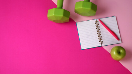 Workout and weight loss equipment. Green dumbbells a notebook a pen and a green apple on a pink...