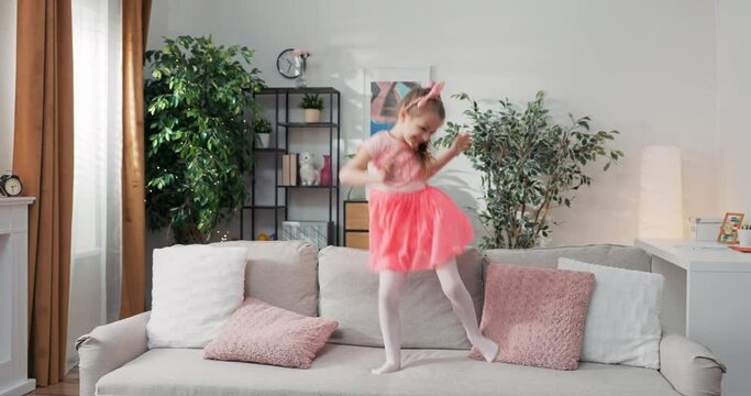 Carefree Childhood. Happy energetic Caucasian girl jumping on the couch while playing at home, young active child.