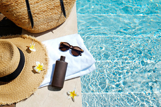 Swimming pool essentials concept. Beach bag with items for safe sunbathing on the deck, sunglasses, straw hat, white blanket & sunscreen product. Flat lay, copy space, top view, background.