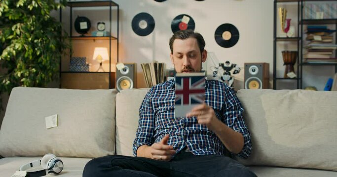 A man tries to memorize new English words. A bearded man sits on the couch and hits head with a dictionary, hoping that he will finally remember the definition.