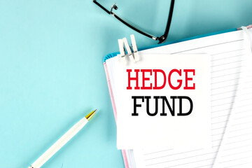 HEDGE FUND text on a sticky on notebook with pen and glasses , blue background