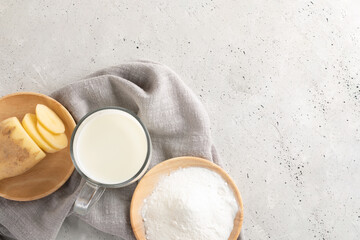 Glass cup with potato milk, potato powder and cut tuber on gray napkin on concrete. Top view.
