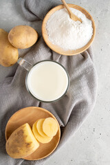 Potato powdered milk in cup, wooden plate with potato powder, tubers on gray napkin on concrete.