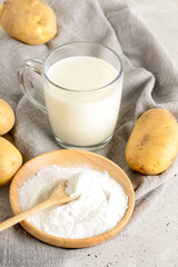 Glass cup with potato milk, potato powder in wooden plate and tubers on gray napkin on concrete.