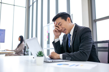 Tired asian businessman with headache at office, feeling sick at work with laptop and book on desk.