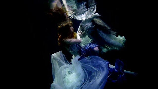 A young girl underwater in the contrasting lighting of the studio by the pool. The smooth fabric of her dress in weightlessness is like in a dream with a dragon.