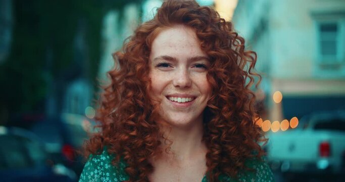 Portrait face young sunshine young woman with red hair look at camera smile stand in the city streets. Beautiful town at sunlight. Feel happy. Slow motion