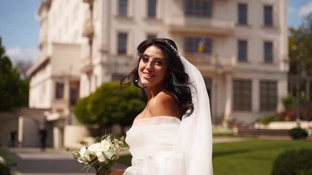 Pretty bride in long airy veil, elegant white dress runs away from camera on wedding day. Playful attractive woman running away turning, looking to viewer. Follow me concept. Fabric fluttering in air.