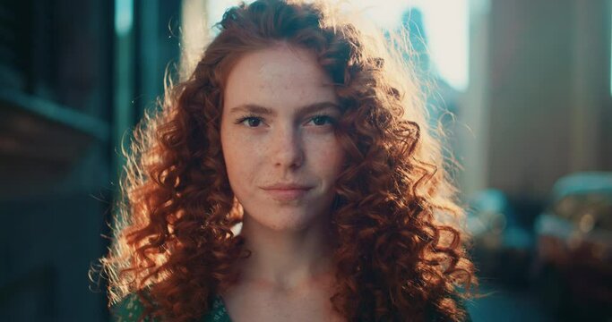 Portrait sunshine young smiling woman with red curly hair look at camera stand in the city streets. Feel happy. Summer. Outdoors at sunlight. Slow motion