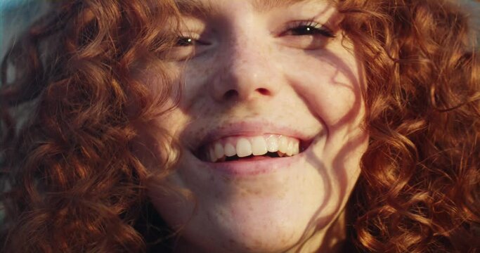 Charming sunshine young smiling woman with red curly hair looking at camera in the city streets at sunlight. Happy portrait face. Slow motion
