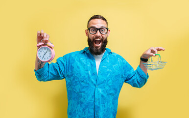 Portrait of smiling charismatic man holding pink alarm clock and mini shopping basket on yellow background. Funny promotion poster. Time to sales