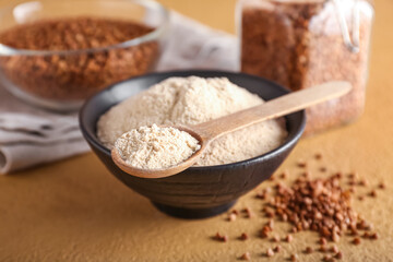 Bowl of flour, spoon and buckwheat grains on beige background, closeup