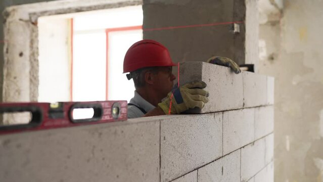 Bricklayer or mason lays bricks to construct wall of autoclaved aerated concrete blocks. Brickwork worker contractor constructs blockwork with foam concrete doing precise masonry with a laser level.