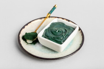 Plate with bowl of spirulina facial mask and powder on light background