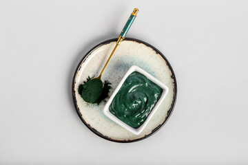 Plate with bowl of spirulina facial mask and powder on light background